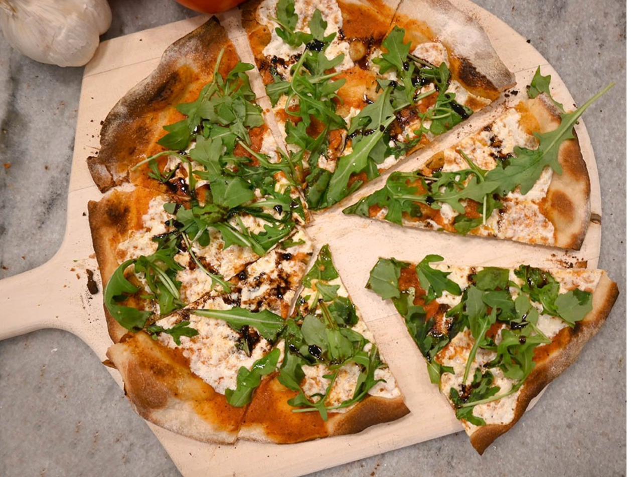 Wood-fired sausage and roasted pepper pizza on a wooden peel, fresh tomatoes and onion behind